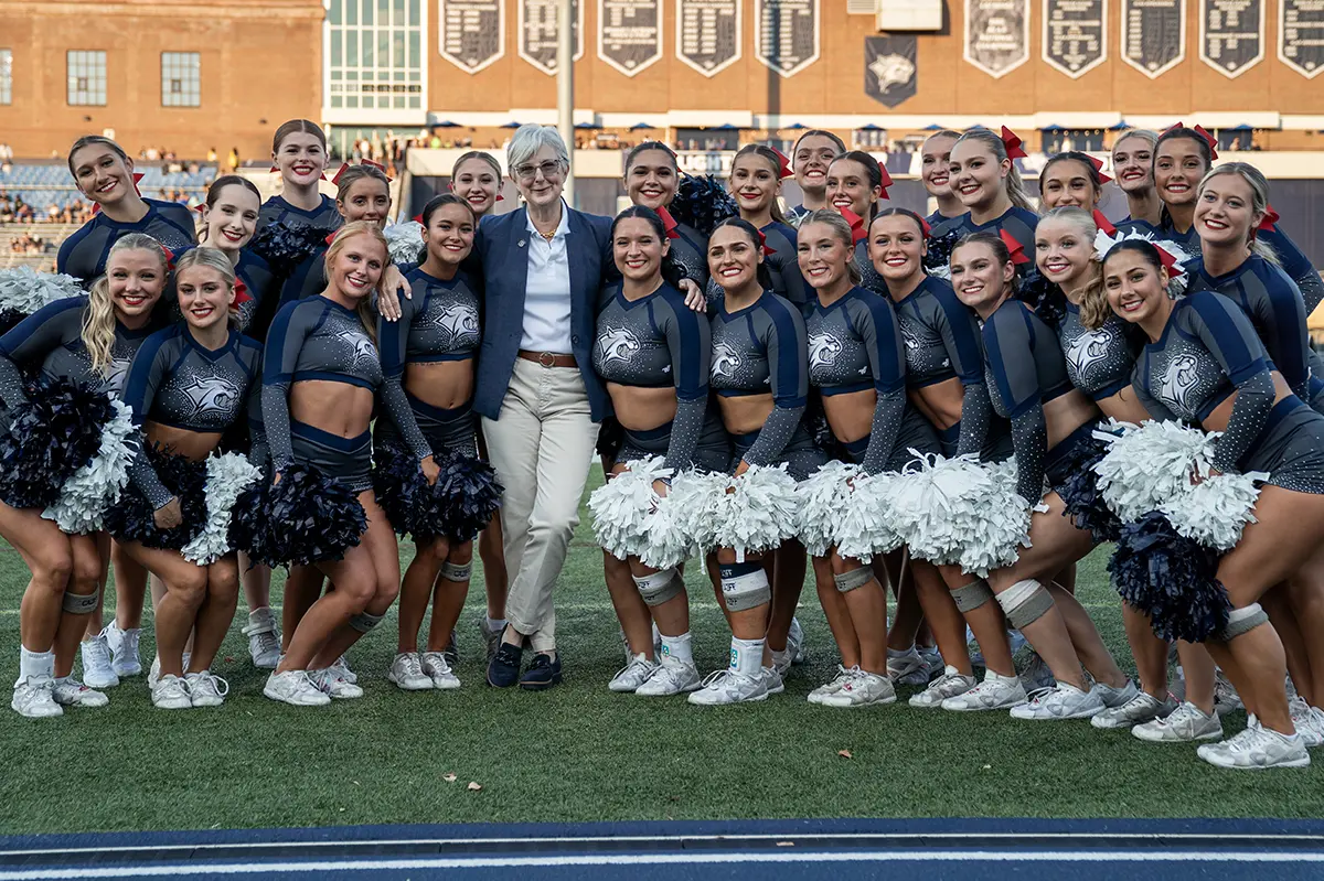 A group of cheerleaders in navy uniforms with pom-poms on a sports field, with a person in a blazer standing with them.