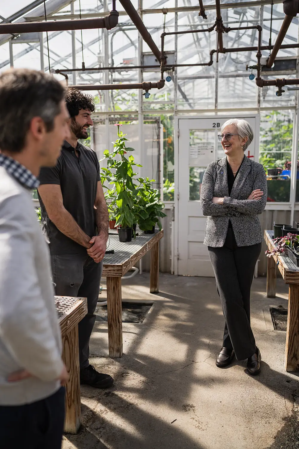 Three people conversing in a greenhouse with potted plants and wooden tables.