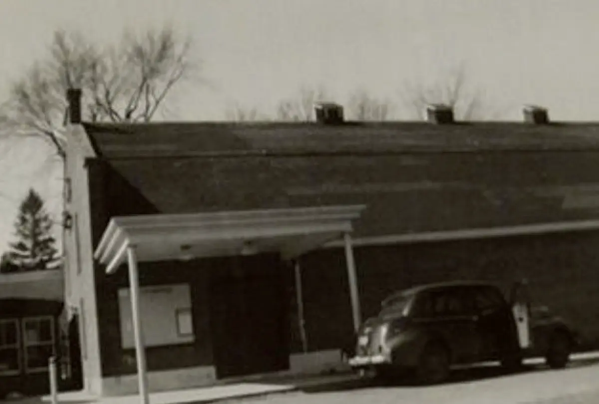 Vintage outdoor black and white photograph showing the exterior of the Franklin Theater on Durham’s Main Street, a small building with a sloped roof; A classic car from the late 1940s is parked in front of a covered entrance; The photo has an aged, sepia tone