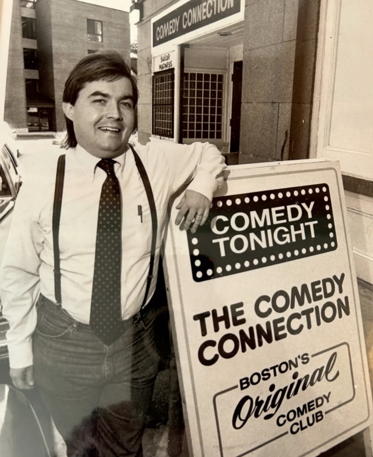 Vintage black and white photo of a man standing outdoors, leaning against a sign that says COMEDY TONIGHT THE COMEDY CONNECTION, BOSTON'S ORIGINAL COMEDY CLUB; The man is smiling and wearing a white button-up dress shirt with suspenders, a tie, and jeans; The entrance to a building is visible in the background; This picture happens to involve Bill Downes ’72