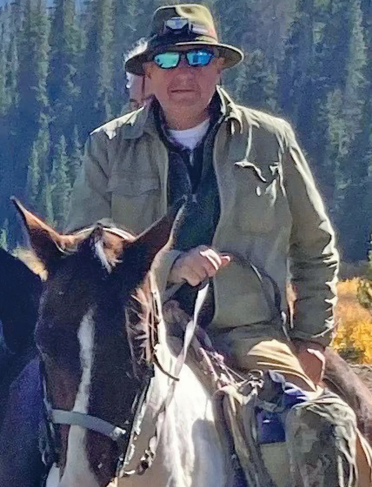 Photo of a man wearing a hat and sunglasses is sitting on horseback, holding the reins; The horse is brown and white; The man is wearing a tan jacket, and a camouflage pack is hanging from the horse's saddle; Trees are visible in the background, with some yellow leaves, suggesting it's autumn; This picture happens to involve Lou Ureneck ’72