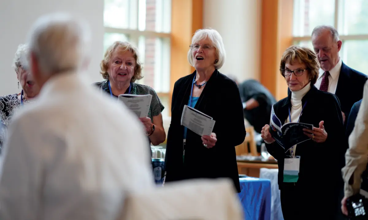 A group of people are standing indoors at an event, appearing to be singing or reading from a booklet; In the center, a woman with white hair and prescription eyeglasses is singing with her mouth open; Others are holding papers and looking at them; A large window and other attendees are visible in the background