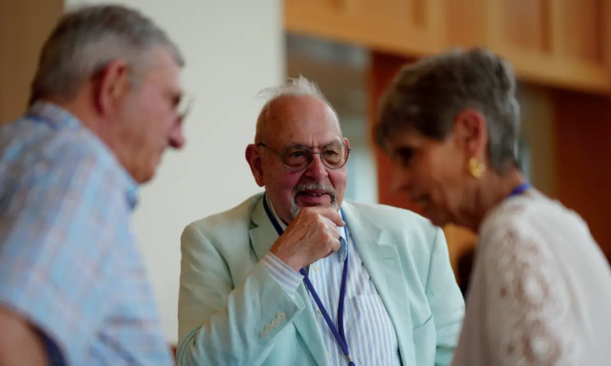 A close-up, candid photo of three older elderly individuals in conversation at an event; The man in the center, with a mustache and prescription eyeglasses, is wearing a light green jacket over a striped shirt and has a conference badge around his neck; He is smiling and looking at the woman on his right, whose back is partially to the camera; Another man is visible on the left, also in conversation