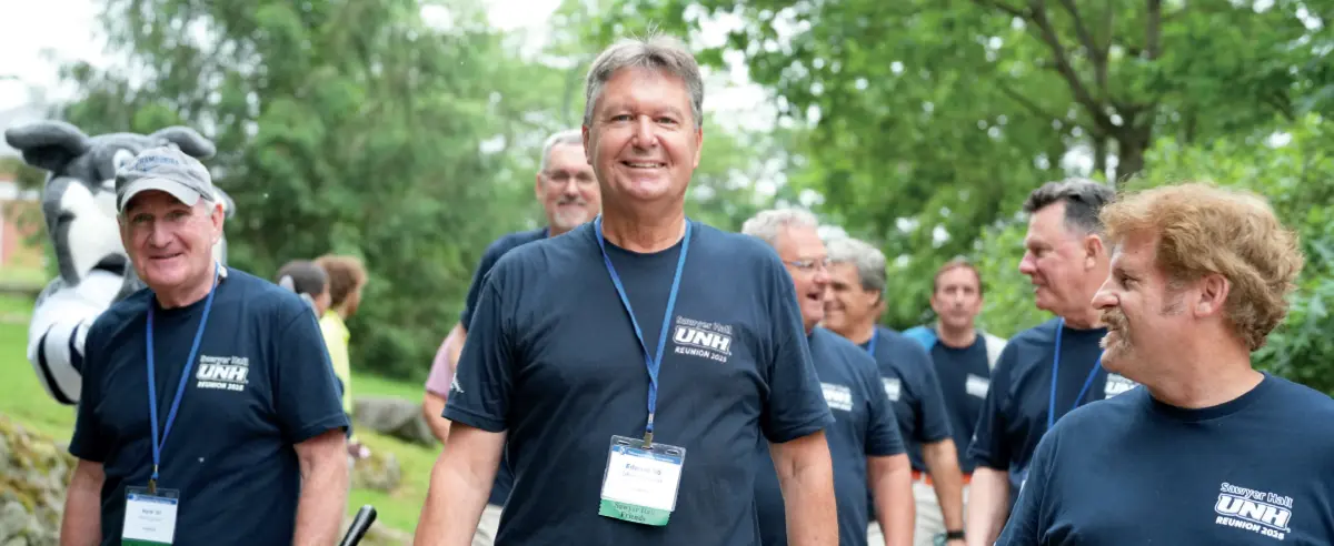 A group of men, most wearing matching dark blue t-shirts that say Semper Uniti UNH, are walking outdoors, smiling and talking; The man in the center is smiling broadly and looking at the camera; A man in a cow mascot costume is partially visible on the far left; The background is a park-like setting with trees