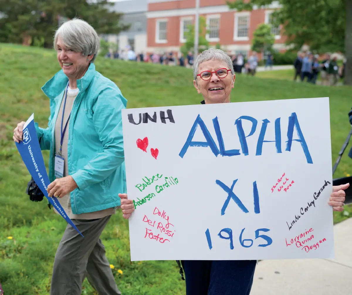 A woman with short gray hair and prescription eyeglasses is smiling at the camera while holding a large white sign that says UNH, ALPHA XI, and 1965 written on it in random various different color phrases; The sign also contains a list of names; She is outdoors on a grassy area with other people in the background as well as an elderly woman nearby her holding a small triangle shaped UNH blue/white banner in her hands and a building in the distance