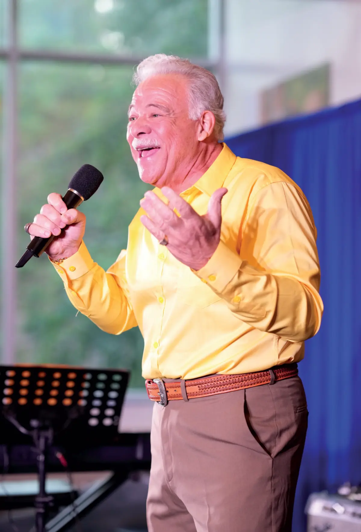 A man with white hair and a mustache, wearing a bright yellow long-sleeved button-up dress shirt and brown pants with a braided leather belt, is singing into a microphone; He is gesturing with his free hand and appears to be performing on a stage with a blue curtain behind him