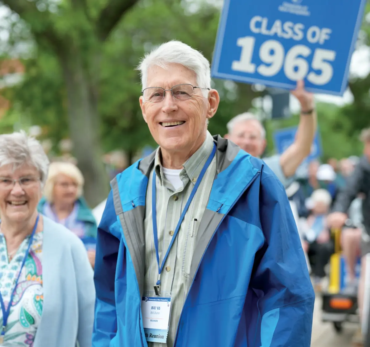 A smiling older man with white hair and prescription eyeglasses, wearing a blue jacket and green pants, is walking in or amongst a group of people outdoors; He has a name tag lanyard badge that says Reunion on it; Behind him, a person is holding a blue sign that reads University of New Hampshire CLASS OF 1965; Another woman with prescription eyeglasses is smiling to his left