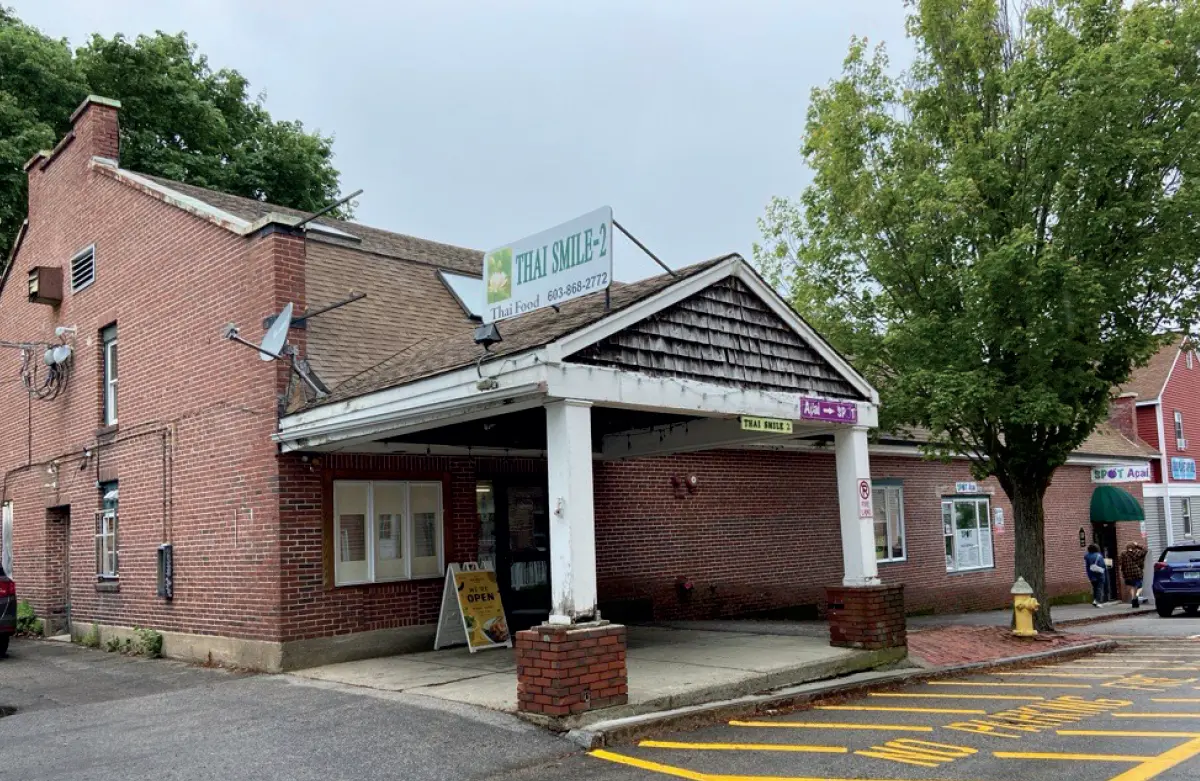 Outdoor photograph showing the exterior of a modern red brick building that houses a business called Thai Smile 2; The building has a prominent covered entryway supported by white columns; The business sign is on the roof, and a sidewalk is visible in front; A yellow fire hydrant, a tree, and a car are also visible in the shot