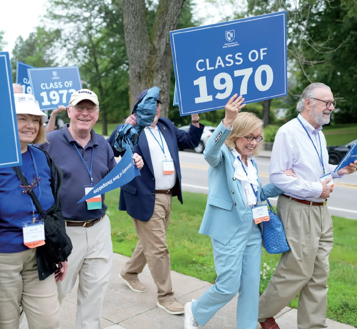 Group of smiling older people are walking outdoors, holding up blue signs that say University of New Hampshire CLASS OF 1970; One woman in a white bucket UNH hat is on the left, and a man in a blue jacket is in the middle with his face obscured by a flag; A woman in a light blue business blazer suit outfit and a man with a beard are on the right walking next to each other holding each other's arms