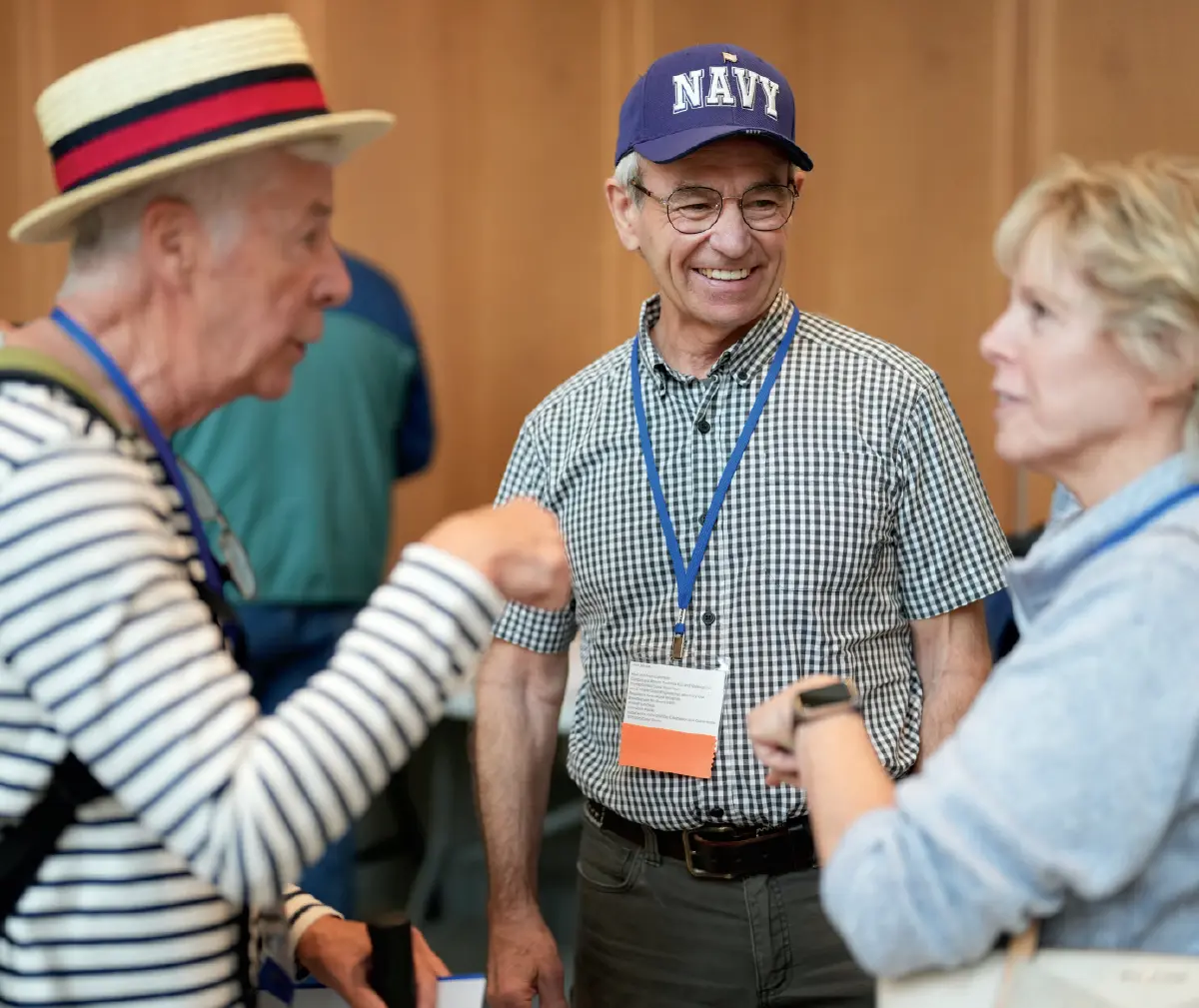 Candid photo of three older elderly people talking at a gathering; The man in the center is smiling while looking at the woman on his right; He wears a blue Navy hat and a checkered button-up dress shirt; The man on the left wears a straw hat and a striped shirt and is gesturing with his fist over at aimed towards the woman in front of him that is pictured