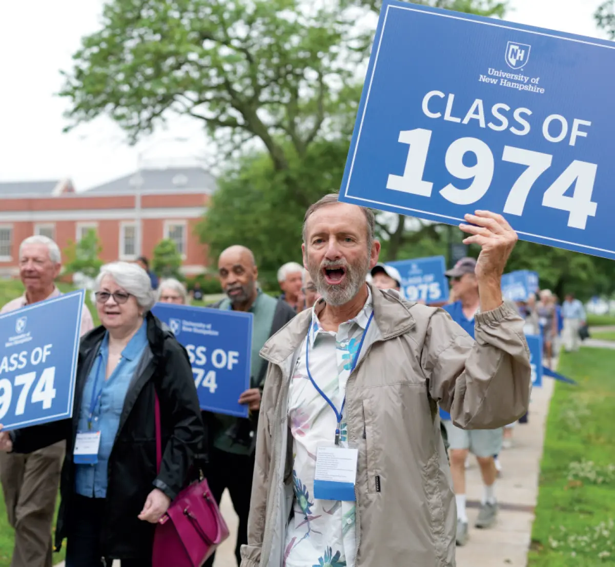 A man with a grey/whitebeard and wearing a dark tan colored jacket is holding a blue sign that says University of New Hampshire CLASS OF 1974 and appears to be cheering or yelling happily; He is walking on a sidewalk with other people around nearby him who are also holding identical same University of New Hampshire CLASS OF 1974 signs; A building and trees are in the background