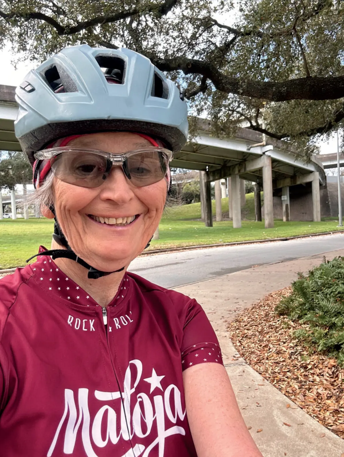 Photo of Laura Rickert Robinson ’74, music education major, a smiling woman with gray hair is taking a selfie outdoors while wearing a bicycle helmet and clear-framed sunglasses; She has on a red jersey with a white design that says Rock 'n' Roll and Maloja; In the background, there is a grassy area and an overpass with concrete pillars