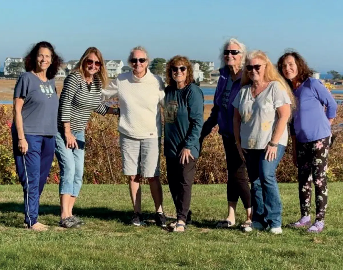 Photo of a group of seven women who are standing together outdoors in a grassy field, smiling at the camera; They are all casually dressed; In the background, there is a body of water with marshy land, and some houses are visible in the distance under a clear blue sky; At their annual UNH gathering, this year in Ogunquit, Maine, are former roomies from the class of ’77 (pictured left to right) Deb Weiss, Mary Ellen Myles, Lauren Chisnall, Wendy Kessler, Jamie Batson, Lauri Hugentobler, Lori Dittelman