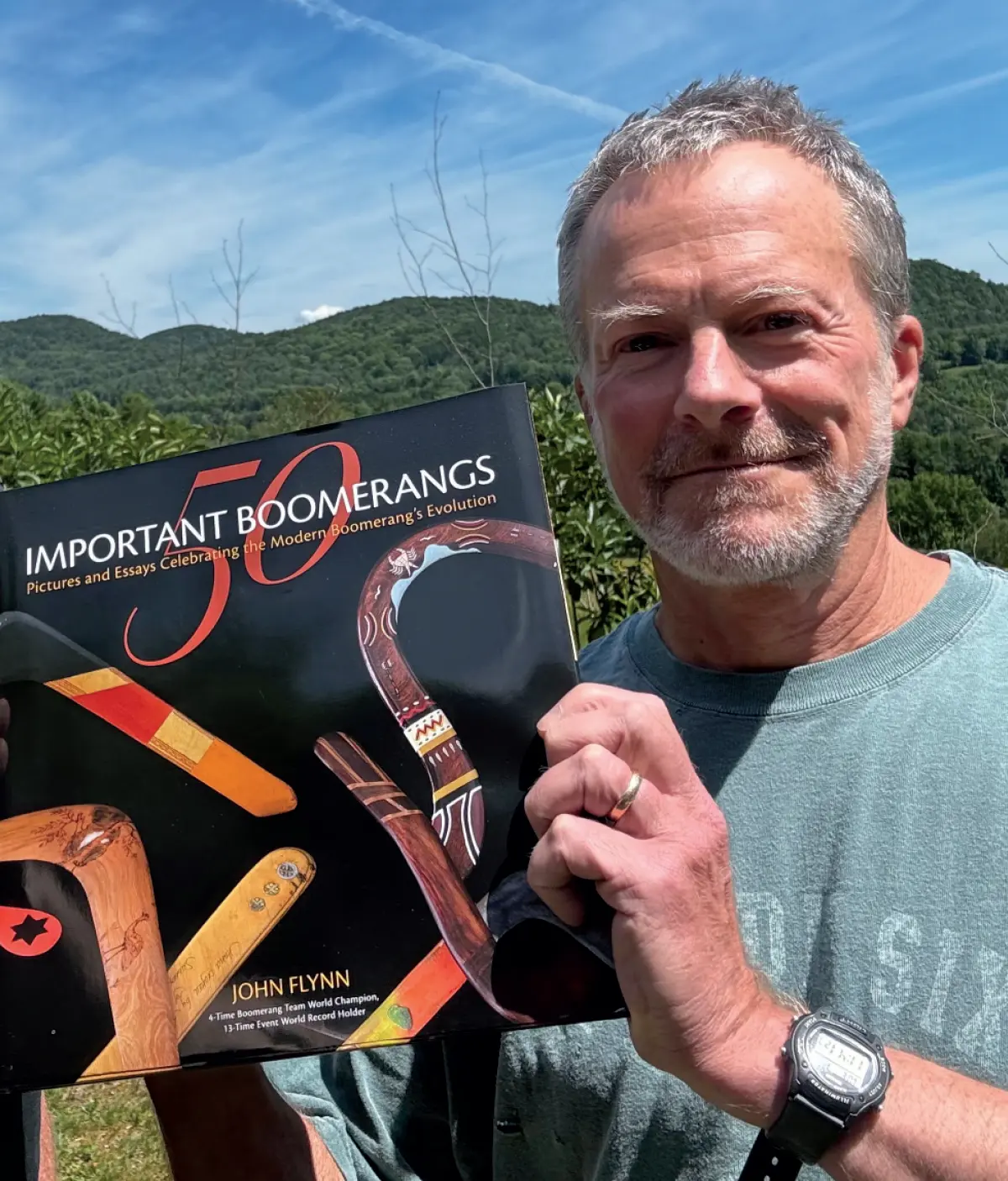 Photo of John Flynn ’80, a grinning man with gray hair and a beard is holding a book titled 50 IMPORTANT BOOMERANGS, Pictures and Essays Celebrating the Modern Boomerang's Evolution; The book's cover features several boomerangs against a black background; The man is wearing a watch and a graphic t-shirt, and he is standing outdoors with mountains and a cloudy sky in the background