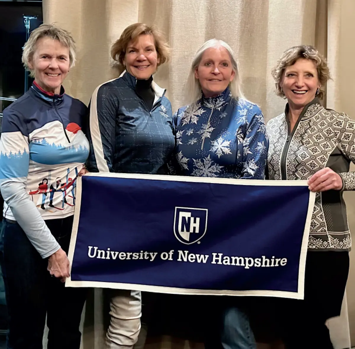 UNH alumni in Trysil, Norway, with the Seacoast Ski Club; Pictured left to right are Debby (Alcox) Mansor ’83, Alexandra Lizak, EMBA ’96, Jen (Roussell) Porter ’81, Caryl Dow ’80; The whole group of individuals pictured here are holding a University of New Hampshire blue/white colored banner flag