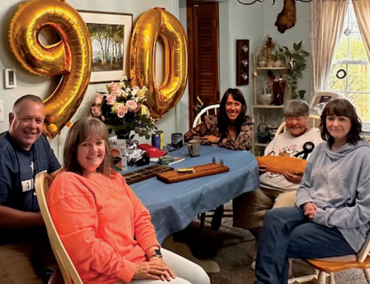 Photo of a group of five adults, four women and one man, are seated around a dining room table celebrating a 90th birthday; A large golden balloon in the shape of 90 hangs on the wall behind them; They are smiling at the camera, and the table is set with a blue tablecloth and a centerpiece of flowers; The room has various home decor items in the background; This happens to be Gwen Marsh Caldwell ’57 and family celebrating