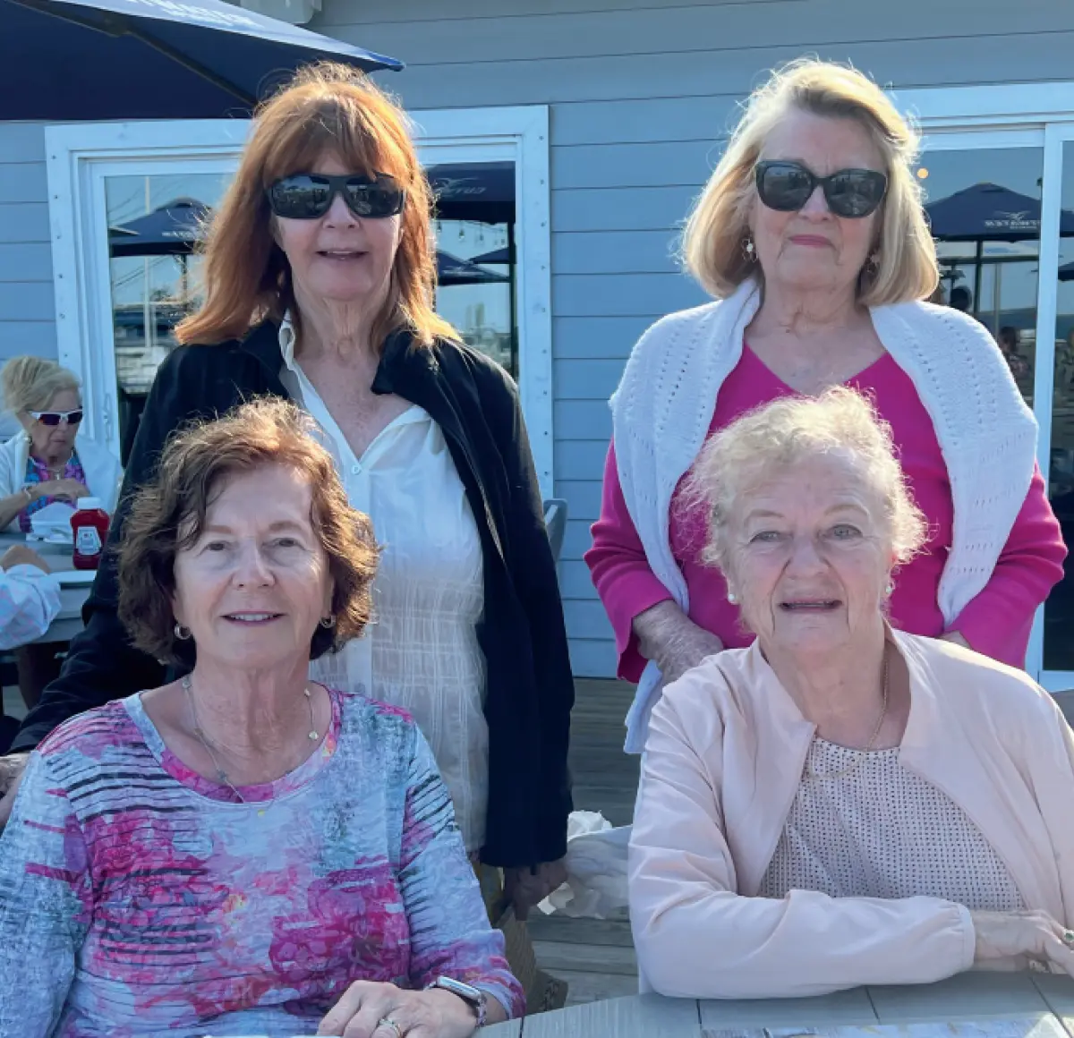 Photo of a group of four smiling older elderly women who are posing for a picture together outdoors on a deck; Two women are sitting at a table in the front, and two women are standing behind them; They are all wearing casual clothing, and two of the women are wearing sunglasses; The background includes a building, tables, and umbrellas, suggesting a restaurant or cafe setting; This picture happens to involve University of New Hampshire Alpha Xi Delta sisters and class of 66ers (clockwise pictured from back left) Linda Kelley Jewett, Sheila Bruce Far, Nancy Perreault Woods and Barbara Starkey Wingardner