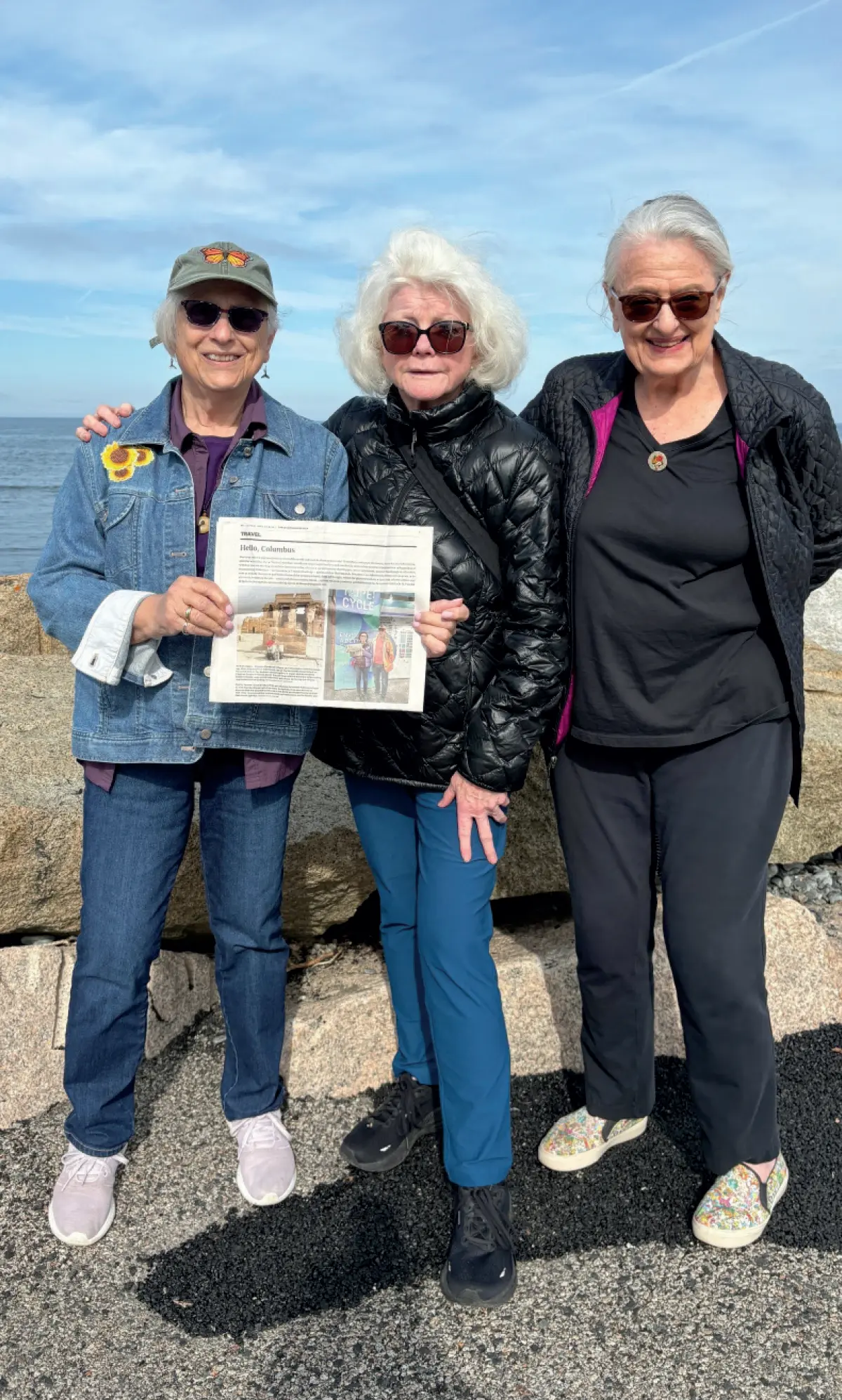 Photo of three women who are standing together outdoors on a rocky shoreline nearby to a body of water under a bright, cloudy sky; The woman in the middle, wearing a black puffer jacket, is holding a newspaper that shows a photo of two people; The women on either side of her are smiling and wearing sunglasses; This picture happens to involve Denise Deschenes ’67, Nancy McGary ’67 and friend Mary O’Connor at Ogunquit’s Marginal Way