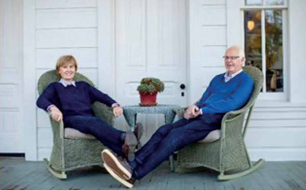 Photo of a man and a woman, both with light hair and wearing matching similar dark blue colored sweaters and pants are sitting in matching wicker rocking chairs on a white porch; They are situated at a small circular shaped round table between them, and both are smiling; The porch has white siding and a large window; This picture happens to involve Lynne Butcher ’69 enjoying retirement in what appears to be her husband sitting across from her