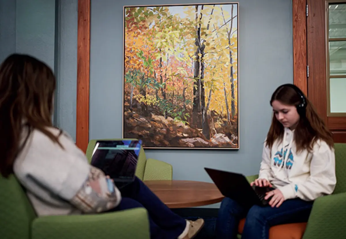 Two university students sit on lounge chairs with their laptops, with a large, colorful painting of a forest scene on the wall behind them.