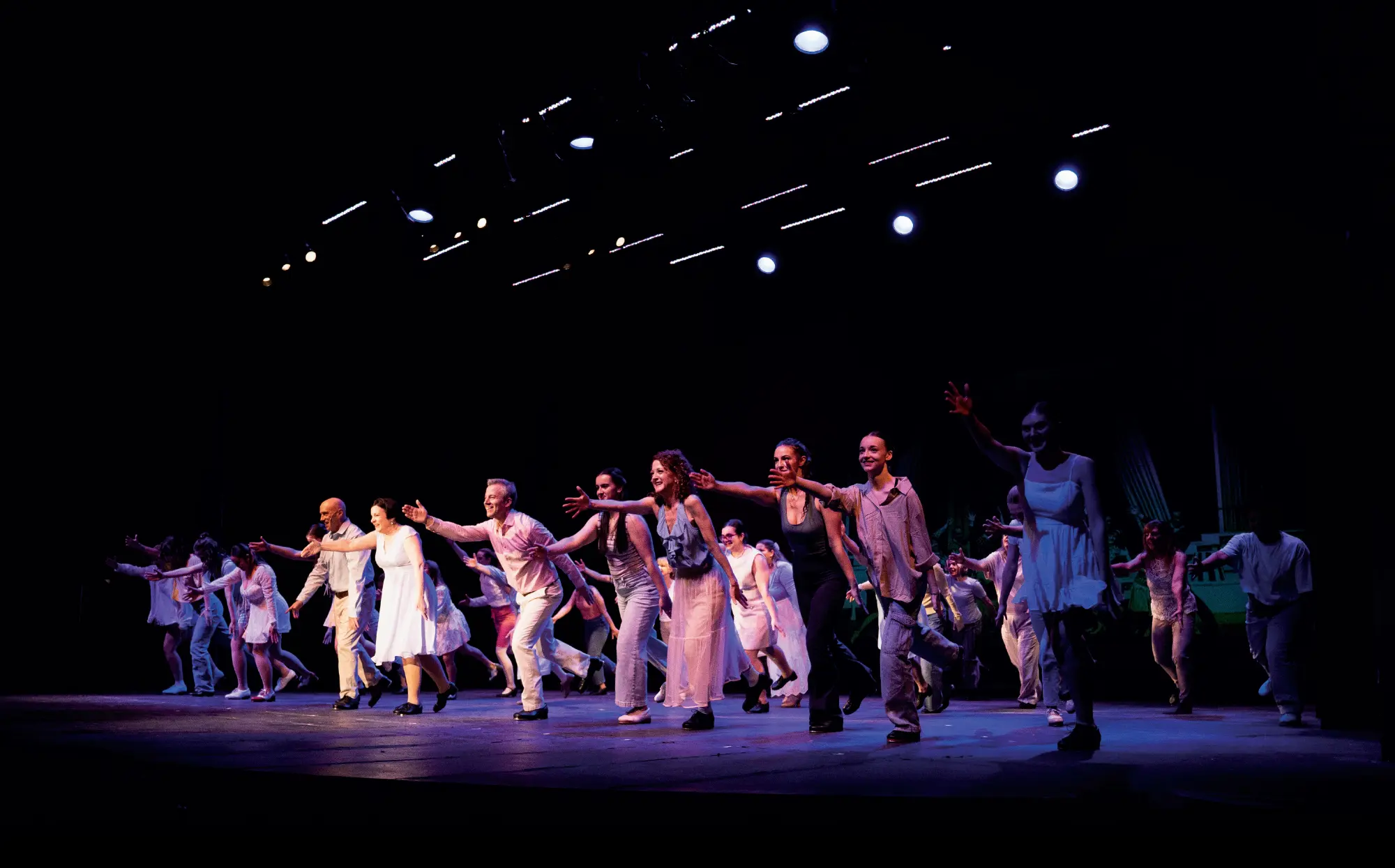 A group of dancers in light-colored costumes performing on a dimly lit stage with a black background and spotlights overhead.
