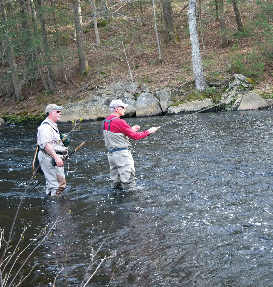 Two men in waders and fishing gear stand in a flowing river, fly-fishing with rods and lines in a wooded area.