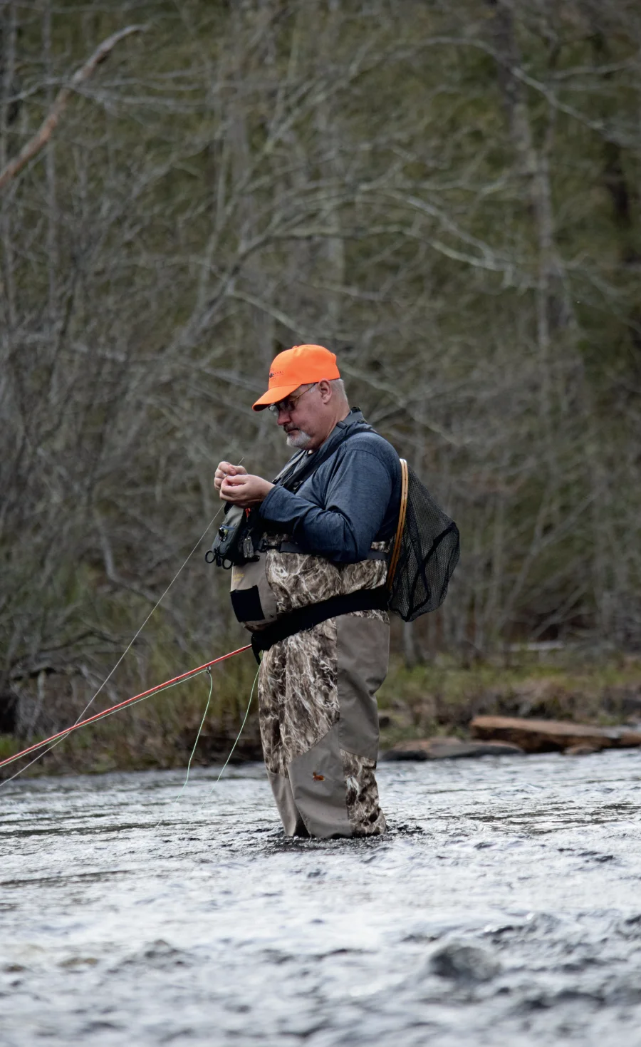 A male angler in waders and an orange cap stands in a river, intently tying a fly onto his fishing line.