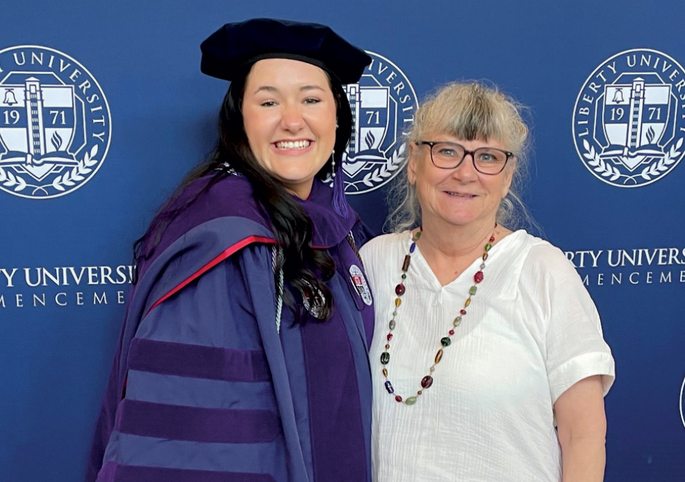 A smiling graduate and an older woman are posing for a picture in front of a blue backdrop with the Liberty University logo. 