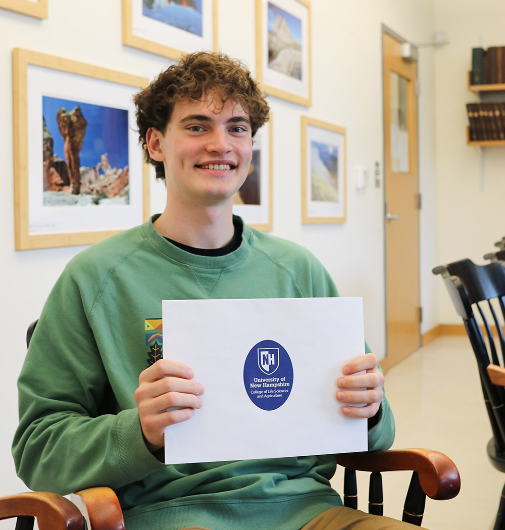 Will Cleveland pictured seated in an office like room, he smiles while holding a UNH labeled folder