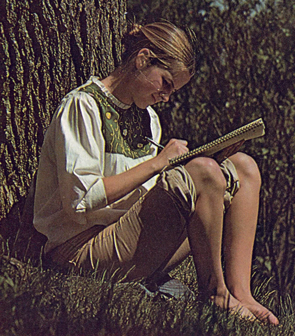 Vintage-style photo of a young woman sitting barefoot against a tree, writing in a notebook outdoors.