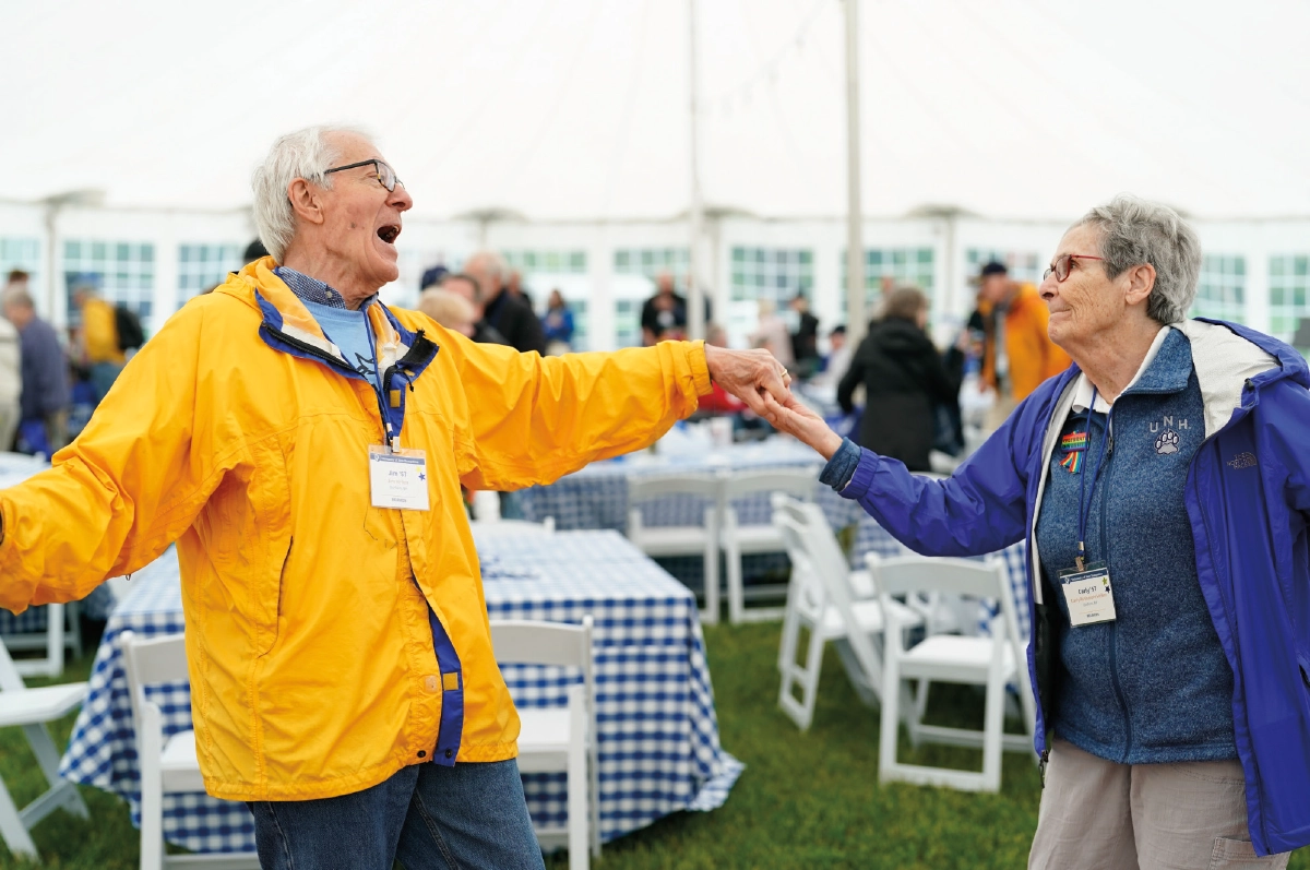 Jim Hellen, an older elderly man in a bright yellow rain jacket and his wife Carly, an older elderly woman in a blue UNH zip-up jacket hold hands and dance joyfully under a large white tent at a recent UNH Reunion event; The man has a wide, open-mouthed expression of laughter
