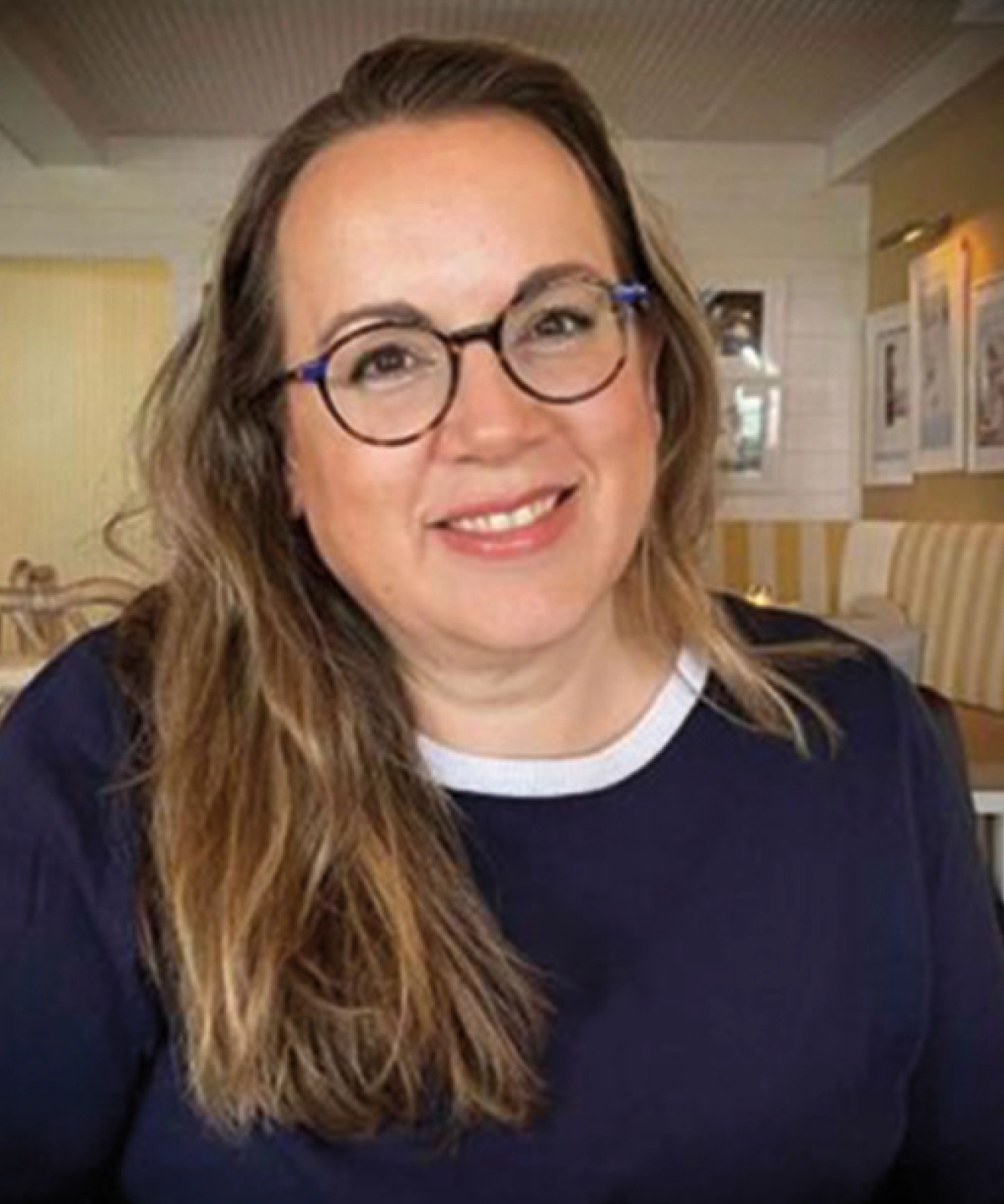 Jennifer Mitchell Perkins, a woman with long, light-brown hair and dark-rimmed prescription eyeglasses, smiling warmly in a dimly lit restaurant setting