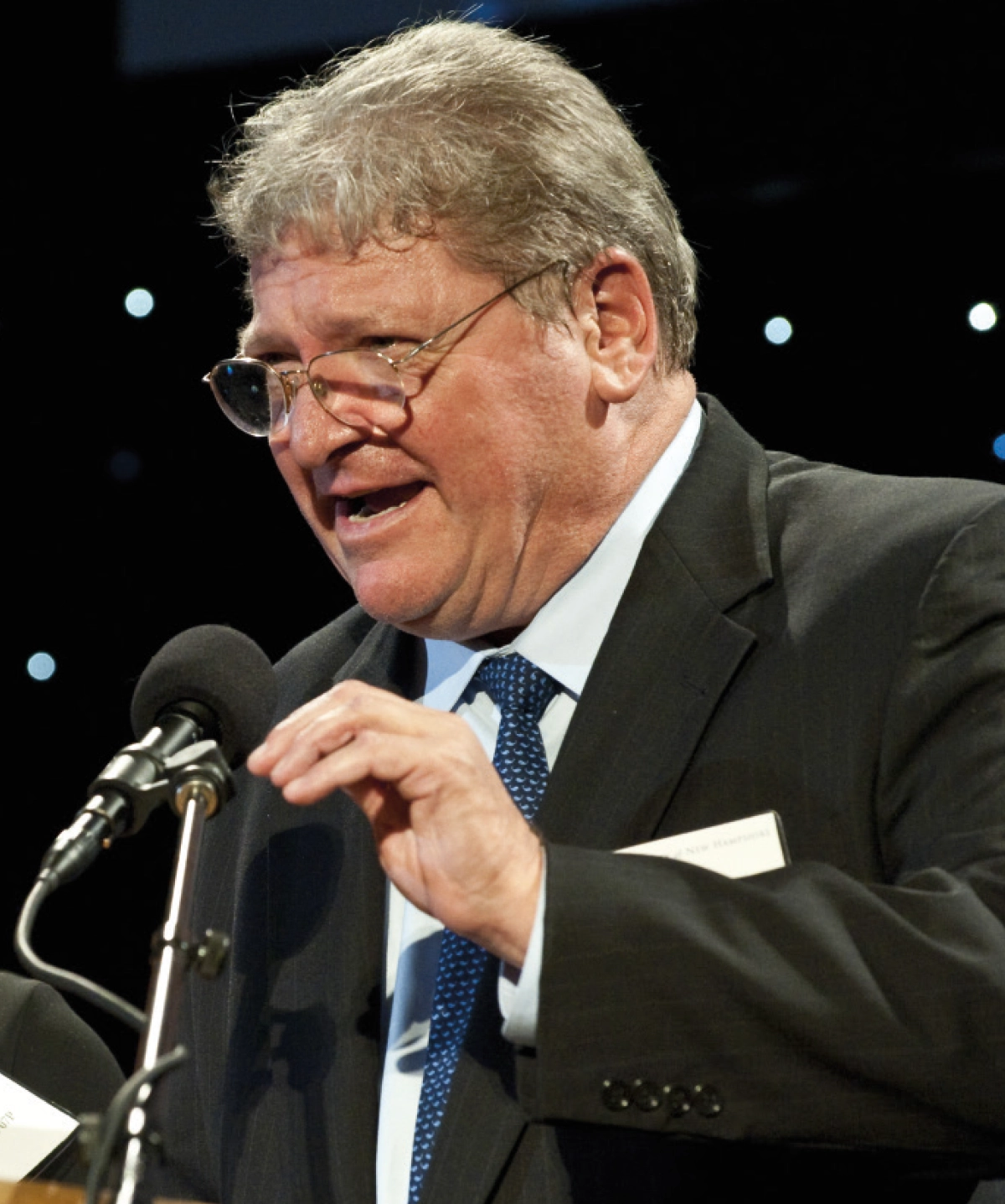 Jim Loomis, an older man in a black suit and prescription eyeglasses speaks into a microphone at a wooden podium against a dark background with light bokeh