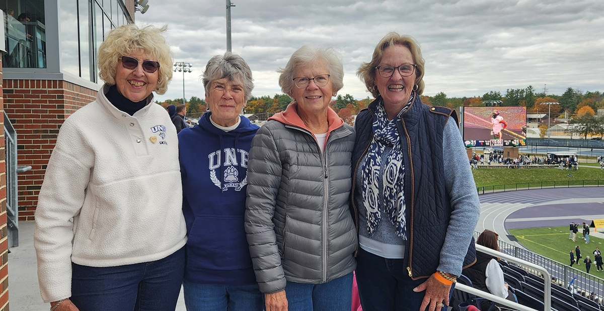Cindy Otis Lindemeyer ’66 had a wonderful UNH weekend in October where she ran into three former classmates (pictured left to right) Kathy Gerbracht Hall, Gail Krippendorf Minschwaner, Gail Hayes Kelly and Cindy Otis Lindemeyer; Here the women are standing together and smiling for a group photo at an outdoor stadium; Two of the women are wearing blue and white UNH apparel