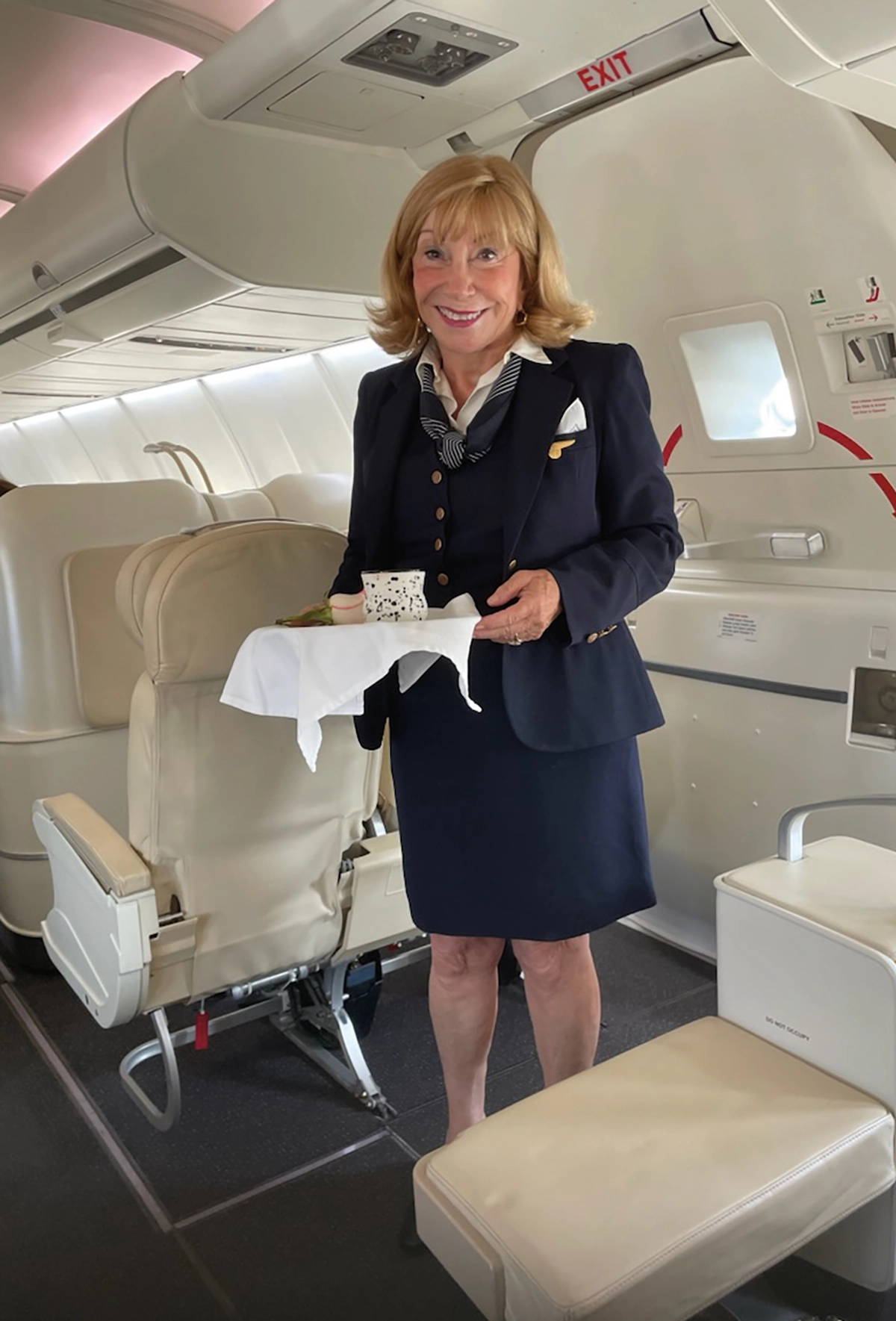 Debbi Martin Fuller ’71, an older elderly flight attendant woman smiling in her own Pan Am navy uniform while on the Tracing the Atlantic flying boat trip of a lifetime holding a serving tray inside the airplane's cabin