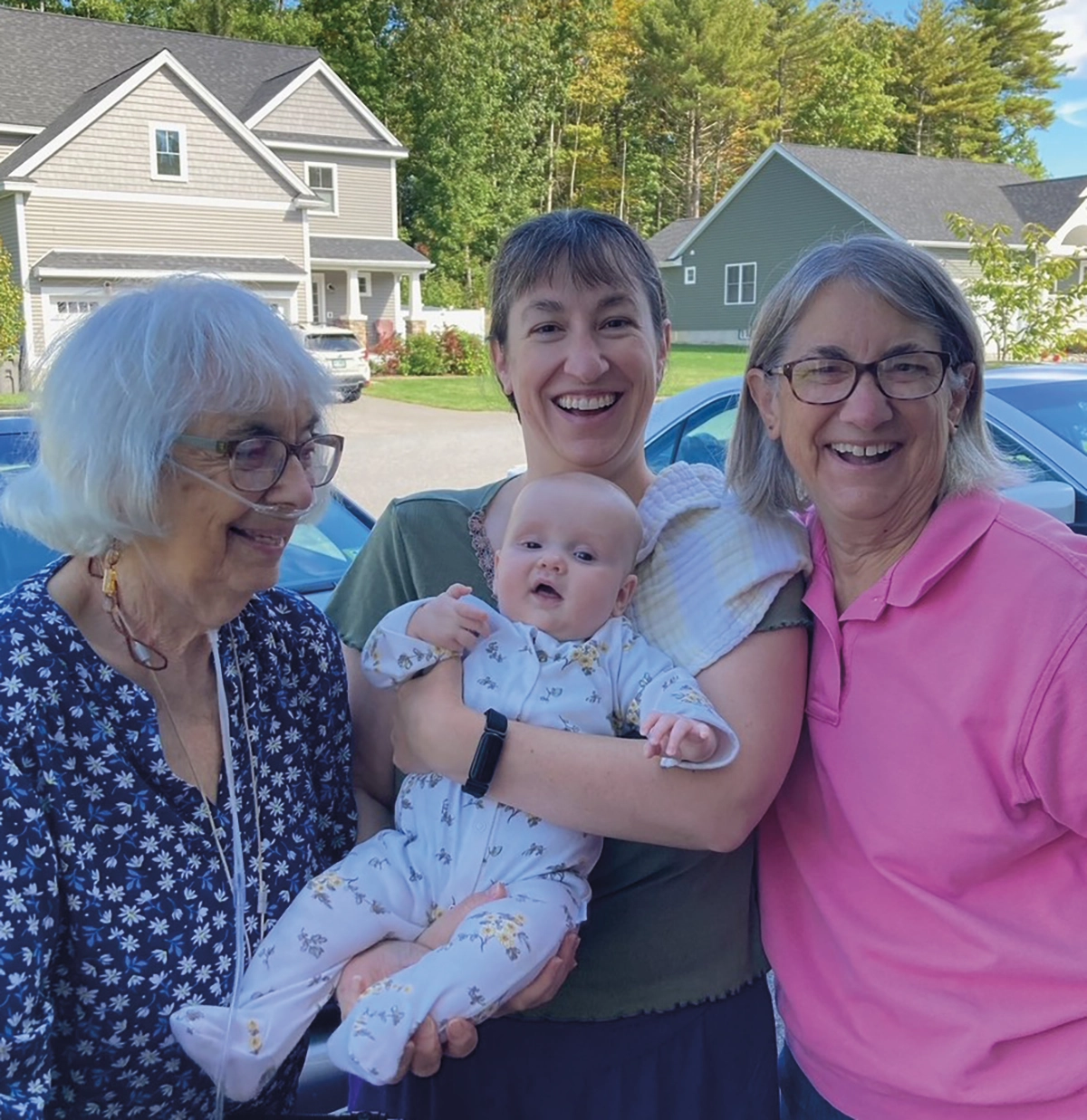 Four generations in Dover: Marilyn Follansbee ’52, Kathryn Burns (Cornell & UNH Shoals program), Lorelie (class of 2046), Vicki Burns ’74 all smiling and posing for a group picture together outdoors on the surface of a home garage driveway nearby some cars, other random houses, and trees located behind them; The woman in the middle is holding a baby