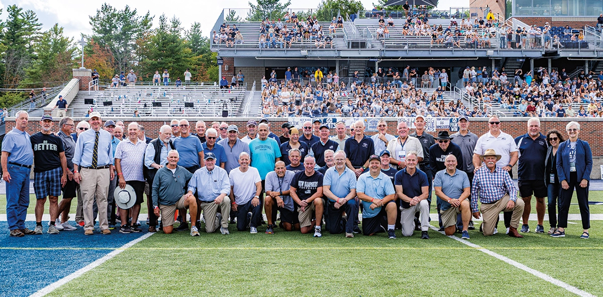 Individuals from the 1975 & 1976 UNH football teams celebrated 50 years together in Durham in September 2025; All of these individuals are gathered together here for a group picture on the football grassy field of a football stadium with other random public audience members in the bleacher stands behind them