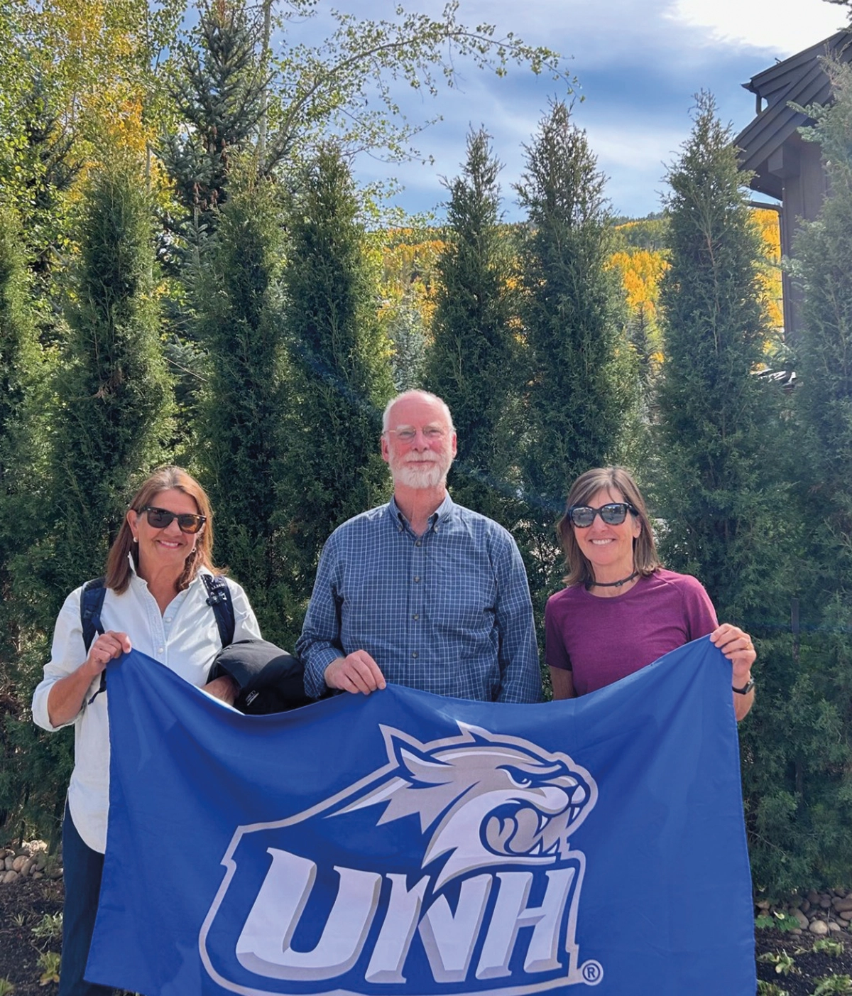 Mona (Fedeli) Stephenson ’79 and Roger Stephenson ’79 with Mona’s cousin Margie Fedeli ’78 in Vail, Colorado as all three of these individuals are standing outside nearby some trees as they are holding a UNH flag of some kind