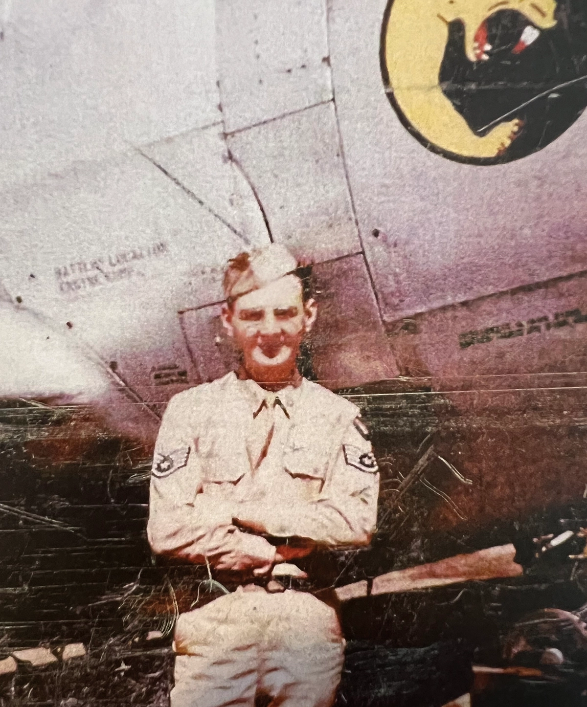 A faded color vintage photograph of Stanley Dabrowski ’56, a man in a light-colored military uniform standing with his arms crossed in front of a fighter jet plane; A yellow circular logo with a black animal is visible on the aircraft behind him