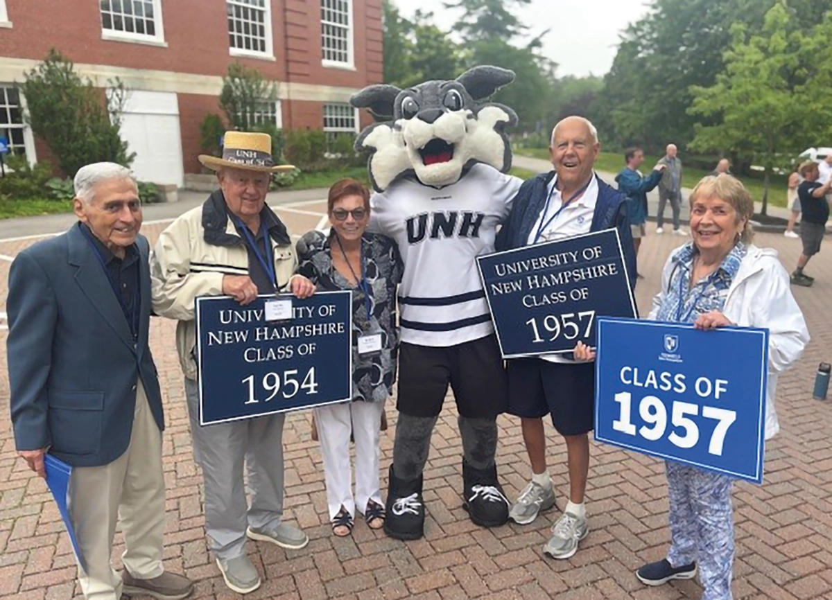 Jim Yakovakis ’57, Lee Perkins ’55, Barbara Sheridan (Fritz’s friend), The Wildcat, Fritz Armstrong ’57, and Nancy Glowacki ’57 had a great time in Durham on UNH Reunion Weekend, June 2025; Here they are all posing for a group picture together outdoors nearby a building of some sort and trees surrounding all around