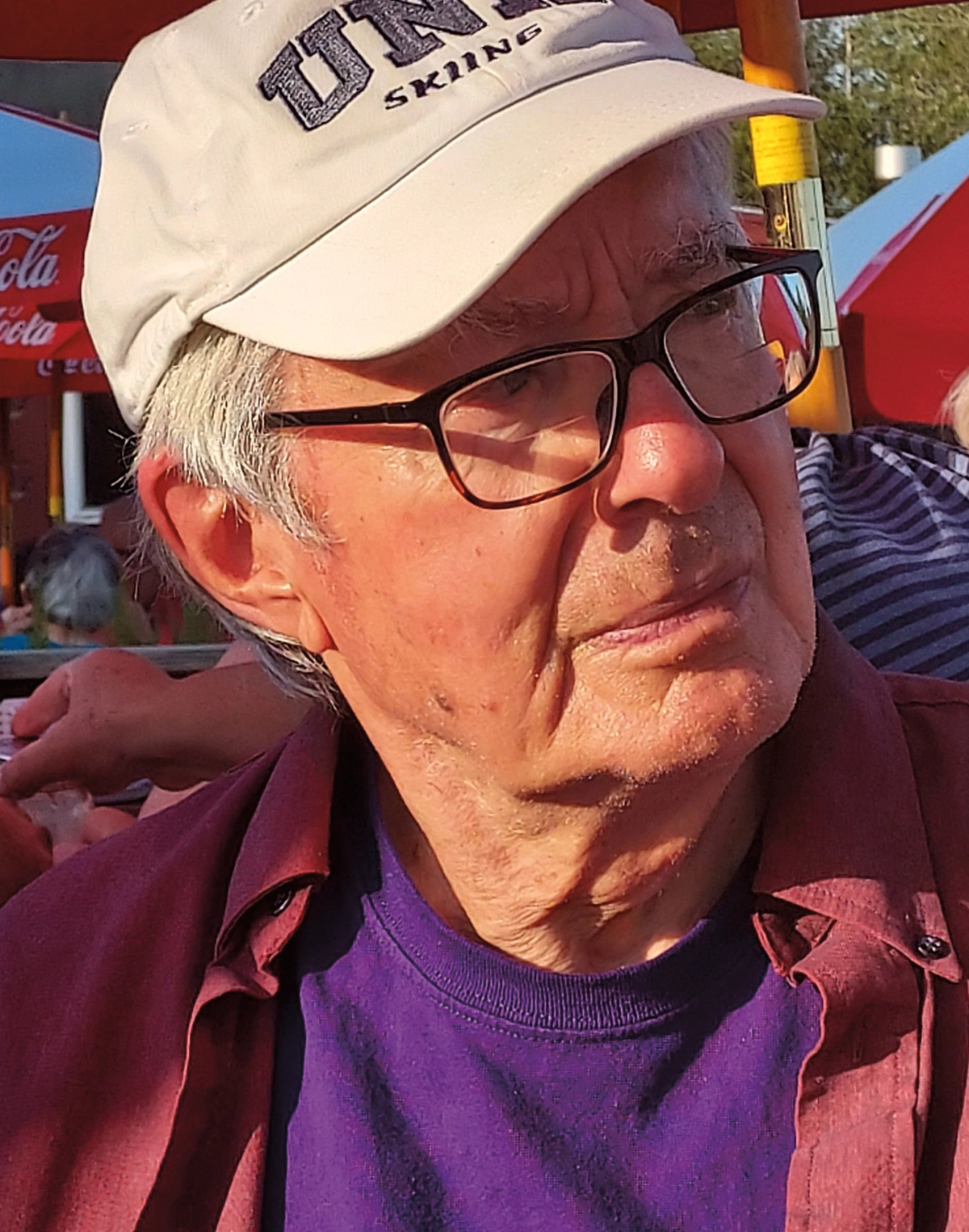 Jim Hellen ’57 in his favorite beige UNH skiing ball cap; Here is he looking away from the camera while wearing prescription eyeglasses, a purple t-shirt, and a maroon open button-up dress shirt