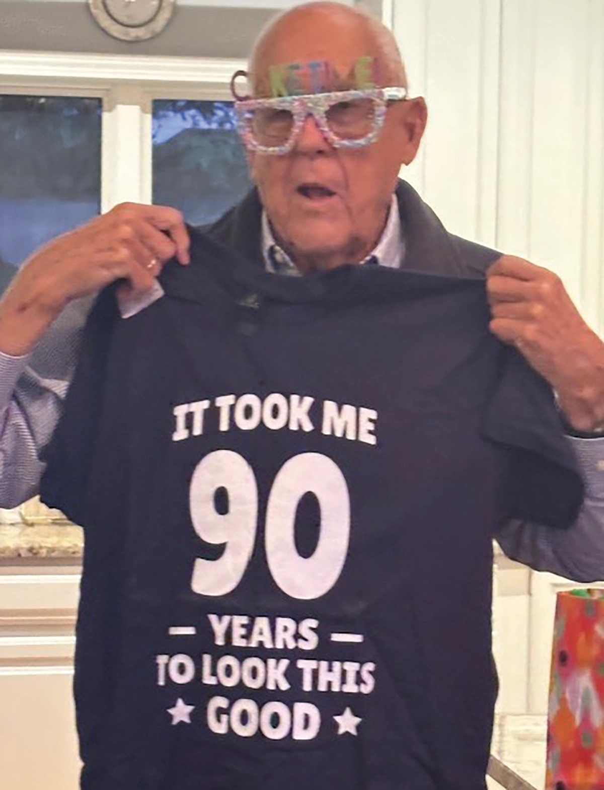 Fritz Armstrong ’57, a joyful older elderly man wearing large, sparkling novelty glasses and a HAPPY BIRTHDAY headband; He is holding up a t-shirt that reads IT TOOK ME 90 YEARS TO LOOK THIS GOOD