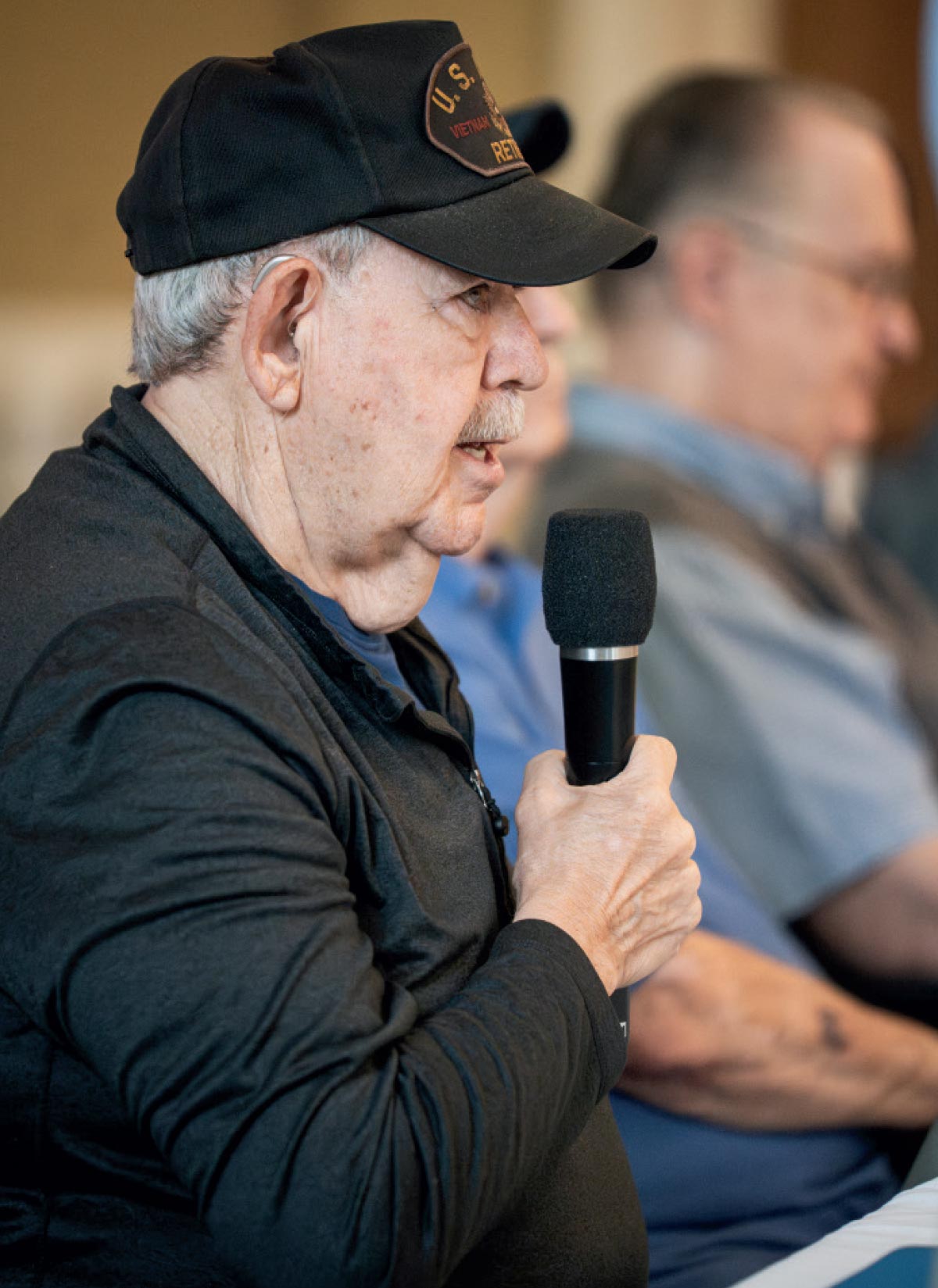 A Vietnam War veteran wearing a retired service cap speaking into a microphone during a community event or panel discussion.