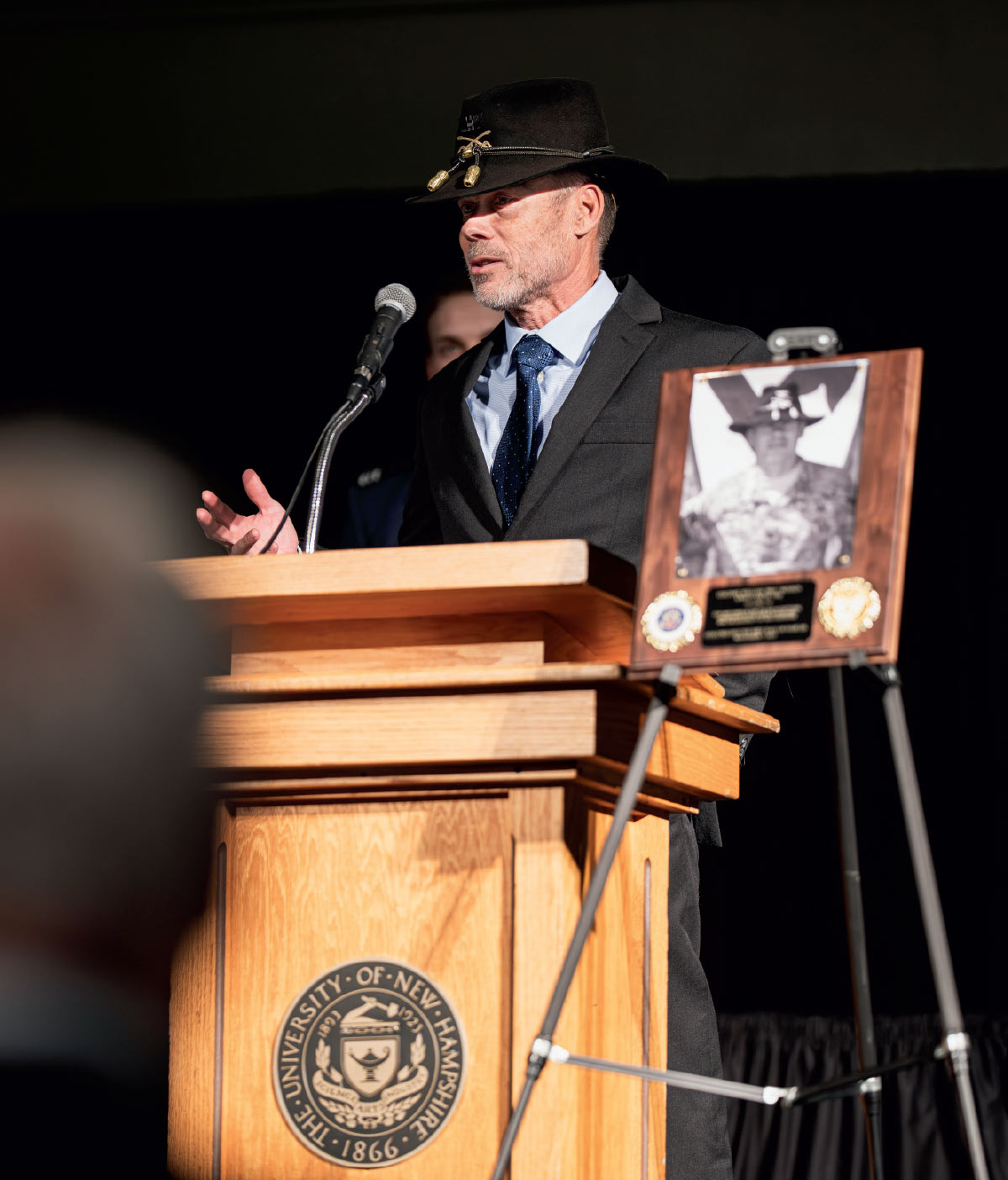 A speaker in a black suit and a military cavalry Stetson hat addressing an audience from a podium at a University of New Hampshire alumni event.