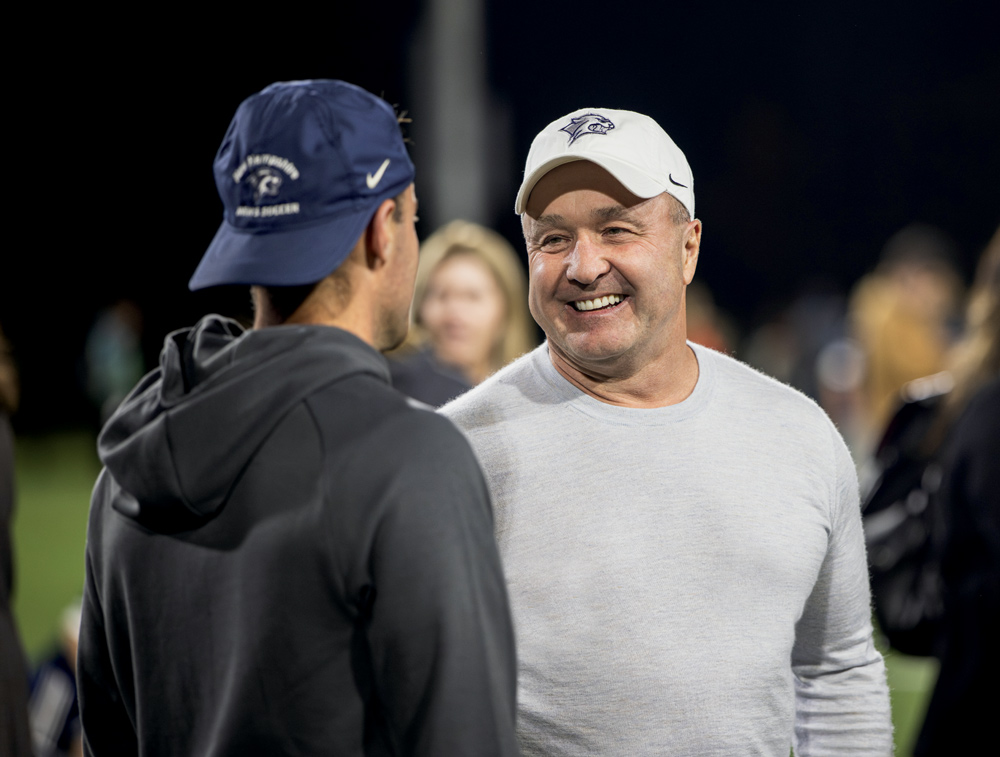 A smiling man in a white baseball cap and light gray long-sleeve shirt faces a man in a navy blue cap and dark hoodie. They appear to be on a sports field at night.
