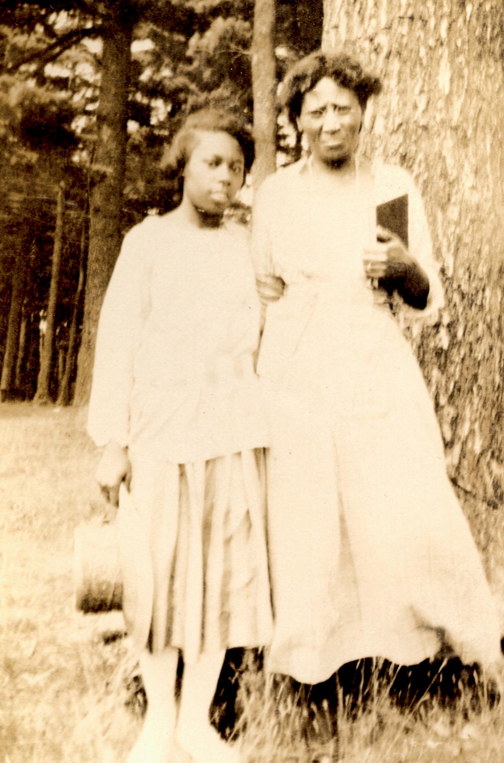 A vintage sepia photograph of two Black women standing outdoors. One woman rests her hand on the other's arm while holding a small book against a large tree trunk.