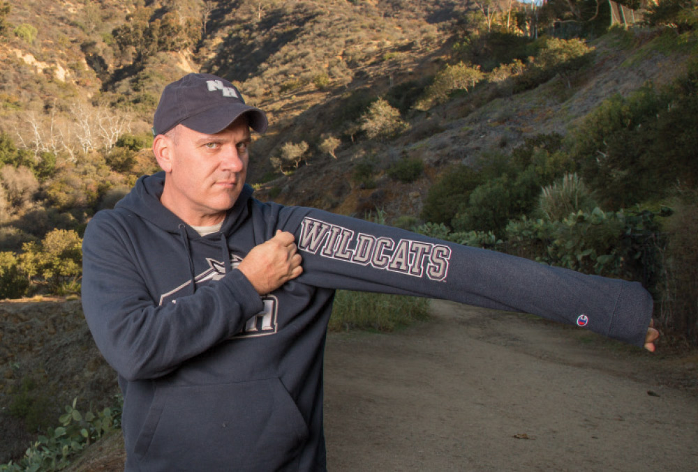 A man in a navy UNH hoodie and baseball cap pulls up his sleeve to display "WILDCATS" printed on the arm.