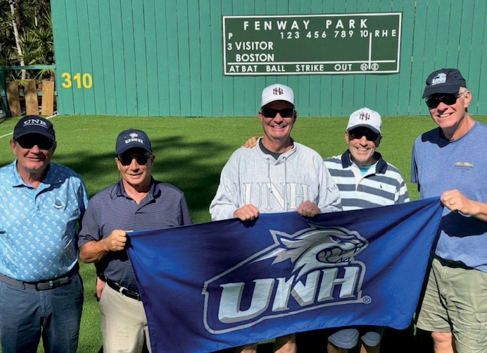 Five men standing in front of a green "Fenway Park" scoreboard while holding a large blue UNH Wildcats flag.