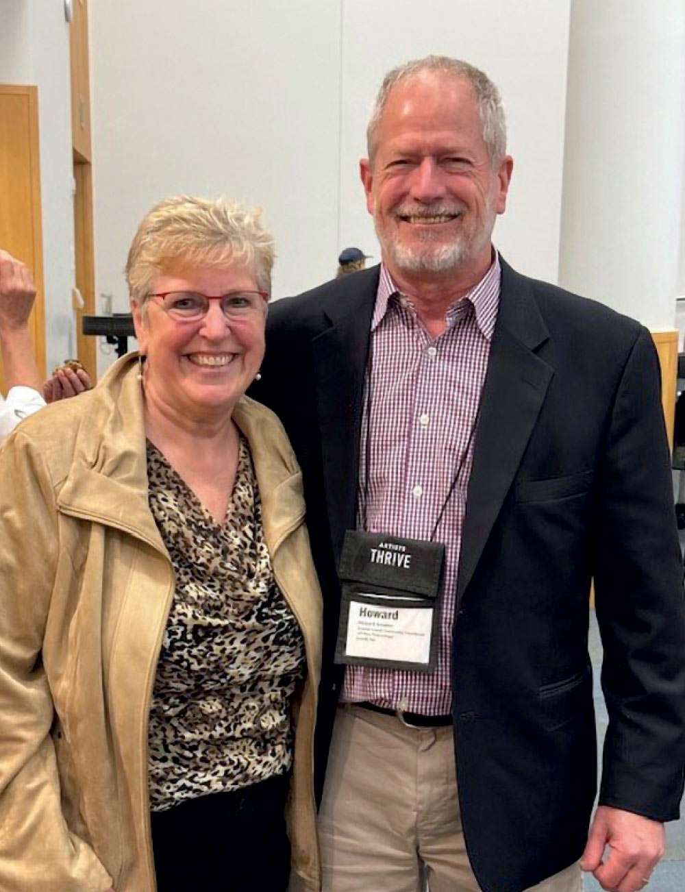 A woman in a leopard-print top and tan jacket smiling alongside a man in a black blazer at an "Artists Thrive" event.
