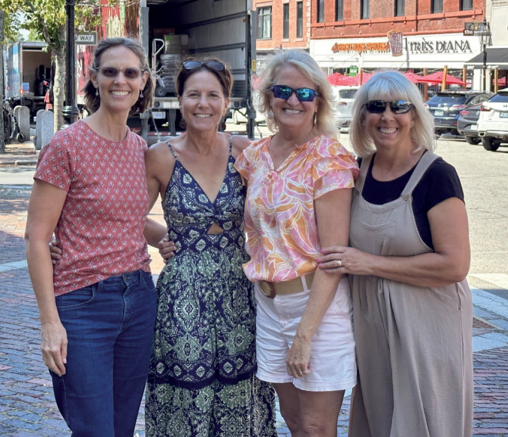 Four women smiling together on a brick-paved city street with storefronts and a delivery truck in the background.